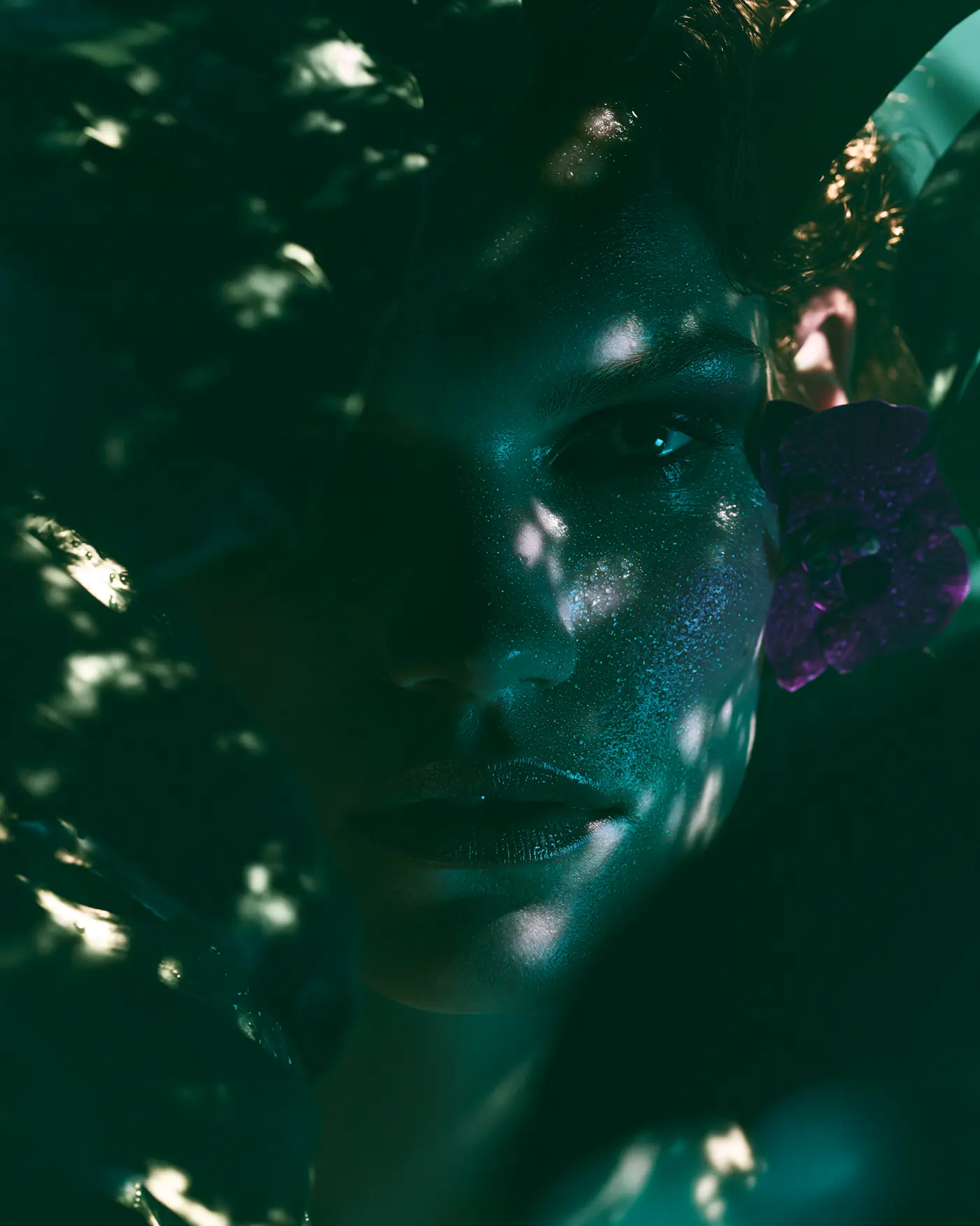 Retrato editorial entre follaje con luz moteada y una orquídea en la oreja
