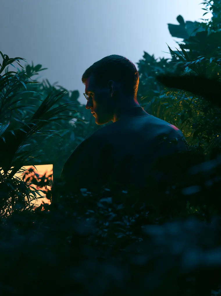 Persona entre plantas tropicales iluminada por el resplandor cálido de una pantalla al anochecer
